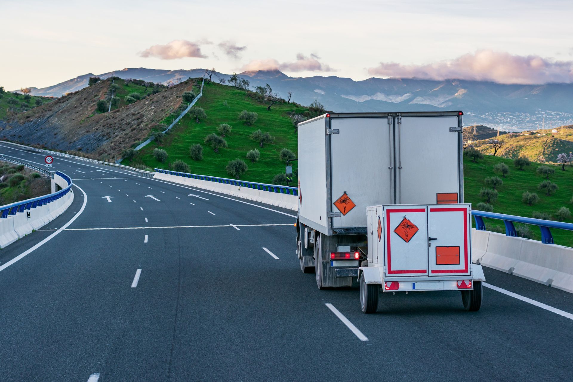 Truck with a trailer carrying hazardous materials drives on a highway with green hills in the background.