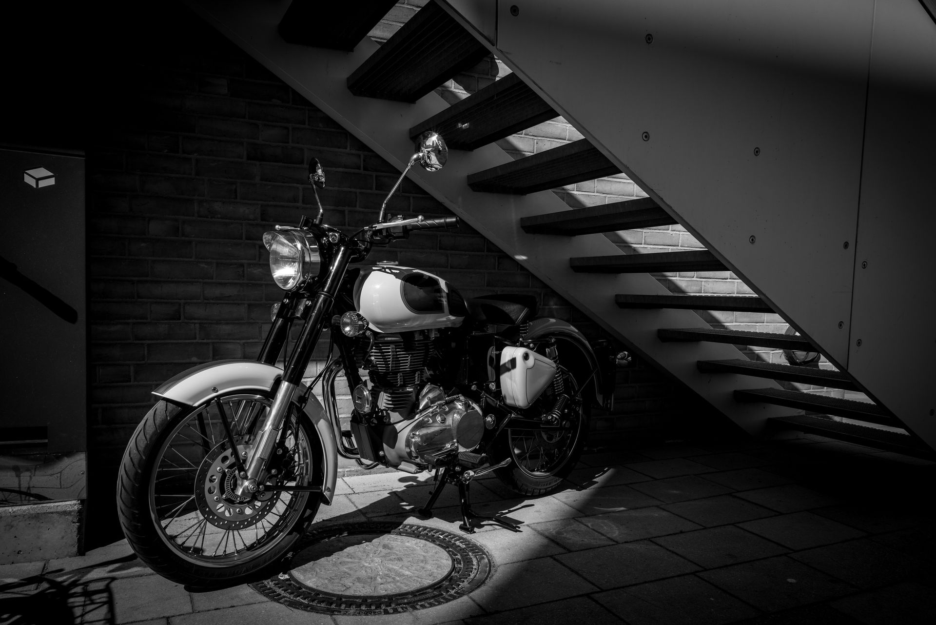 Black and white photo of a motorcycle parked beneath a staircase, illuminated by sunlight.