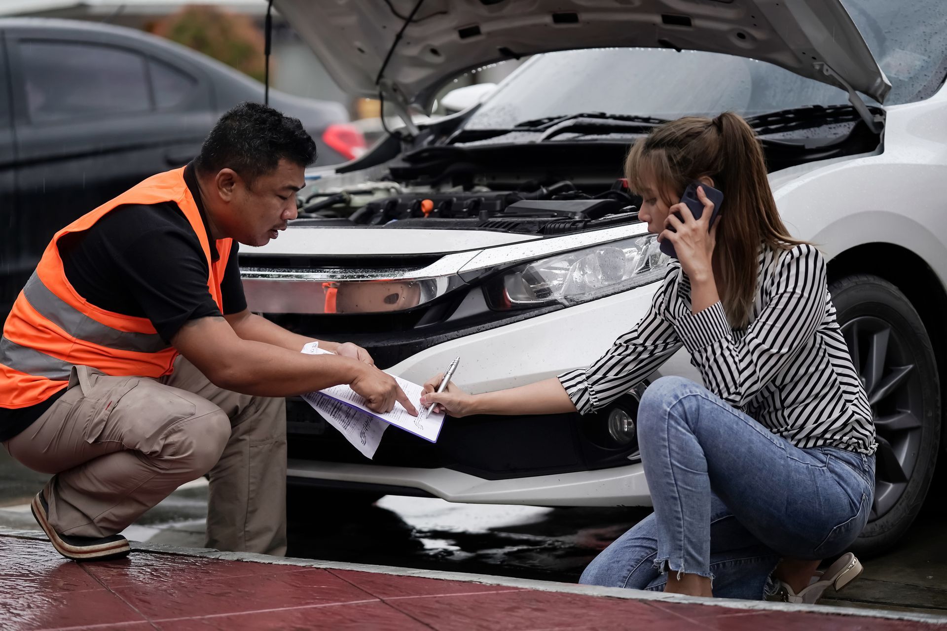 Man and woman by car with open hood, woman on phone signing papers.