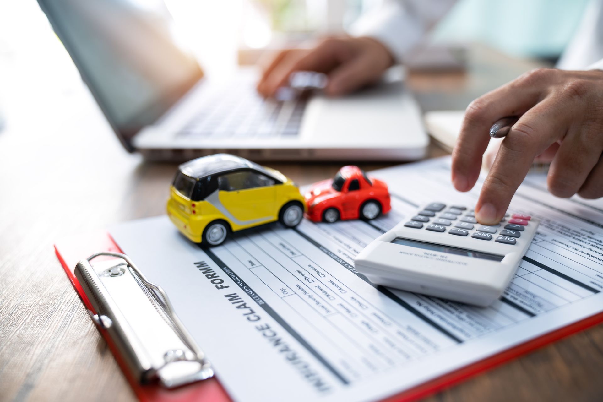 Person calculating insurance on paperwork with toy cars and laptop.