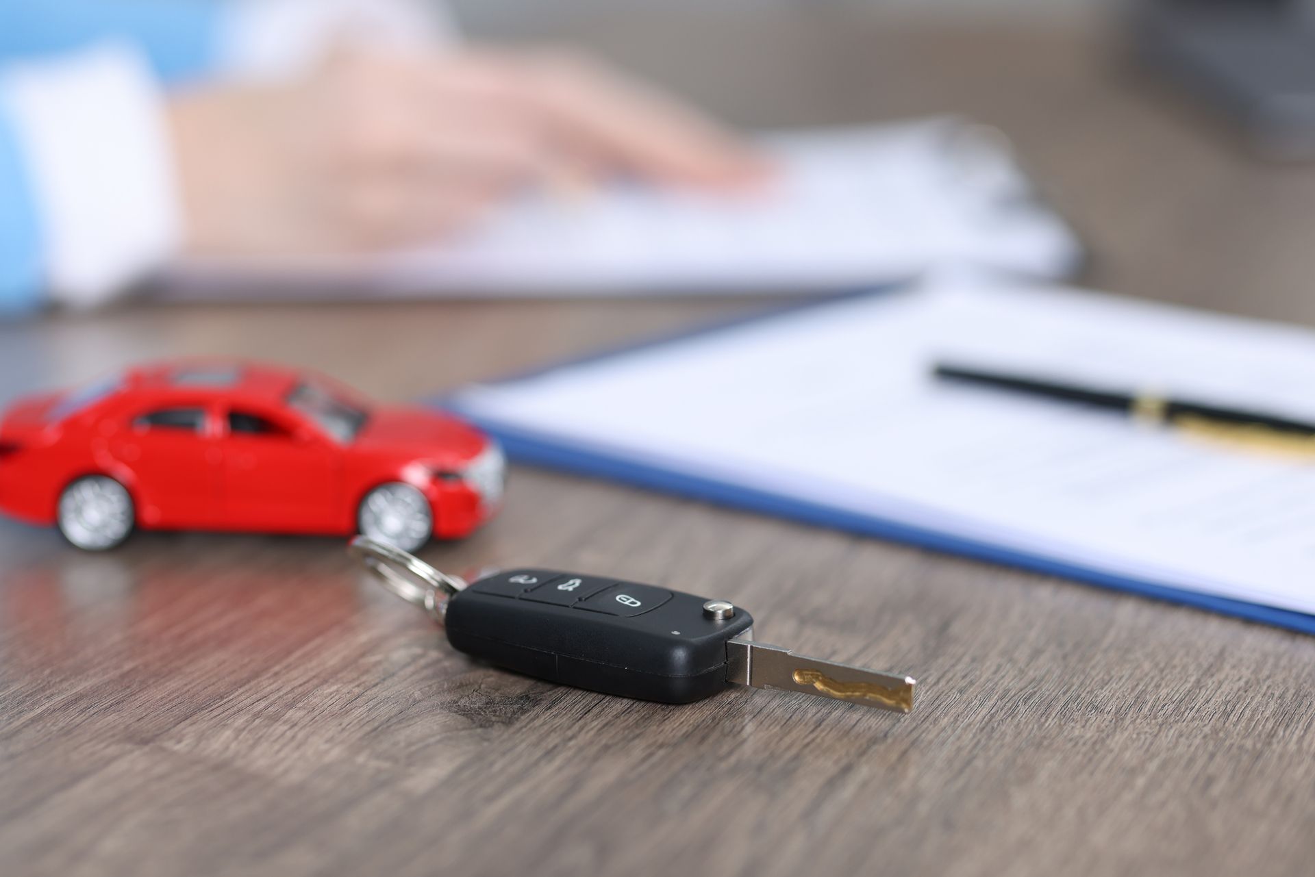 Car keys next to a small red car on a desk near documents and a person working. Car keys next to a small red car on a desk near documents and a person working.