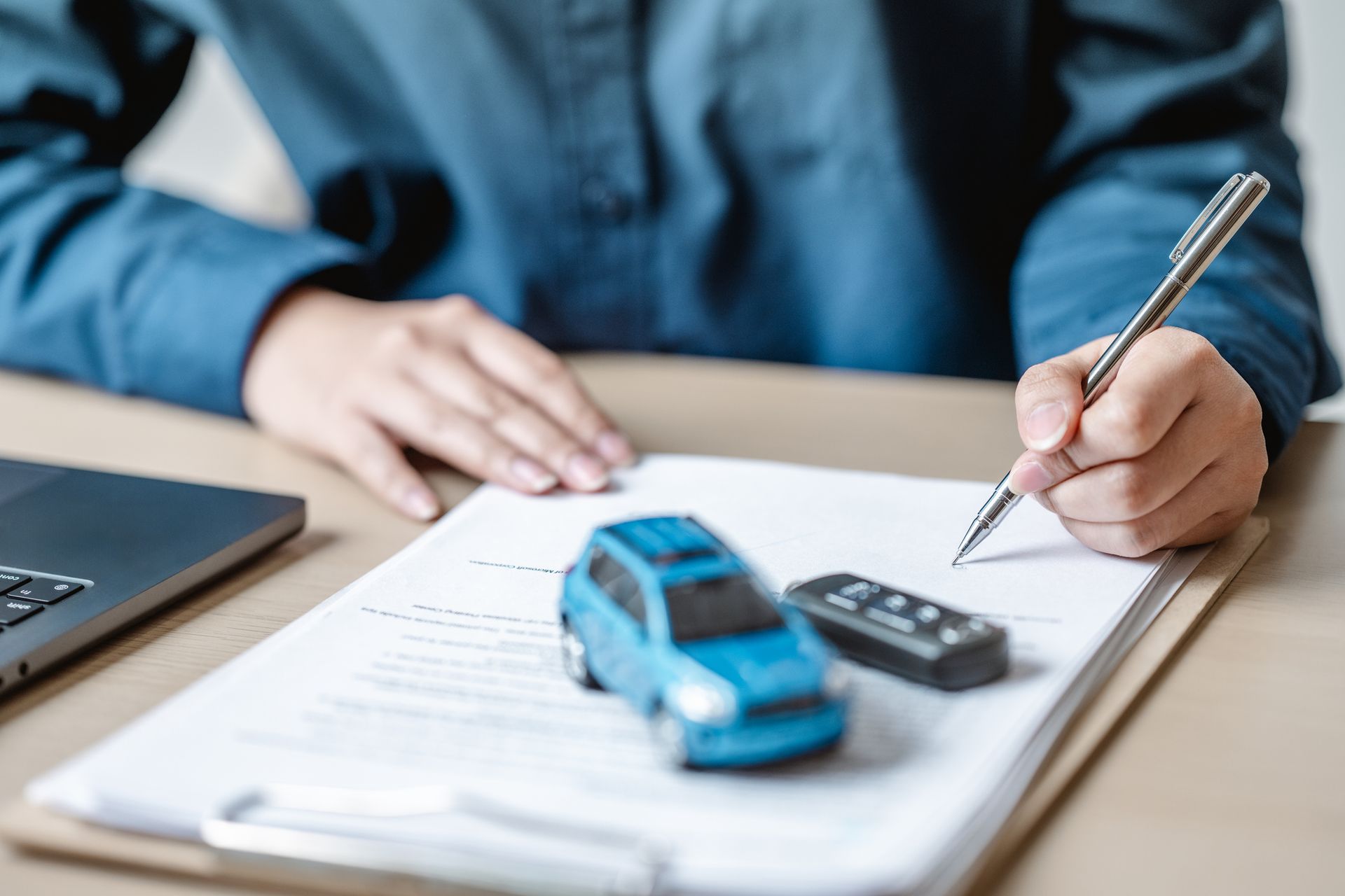 Person signing a document, with a blue toy car and remote on the paper, next to a laptop. Person signing a document, with a blue toy car and remote on the paper, next to a laptop.