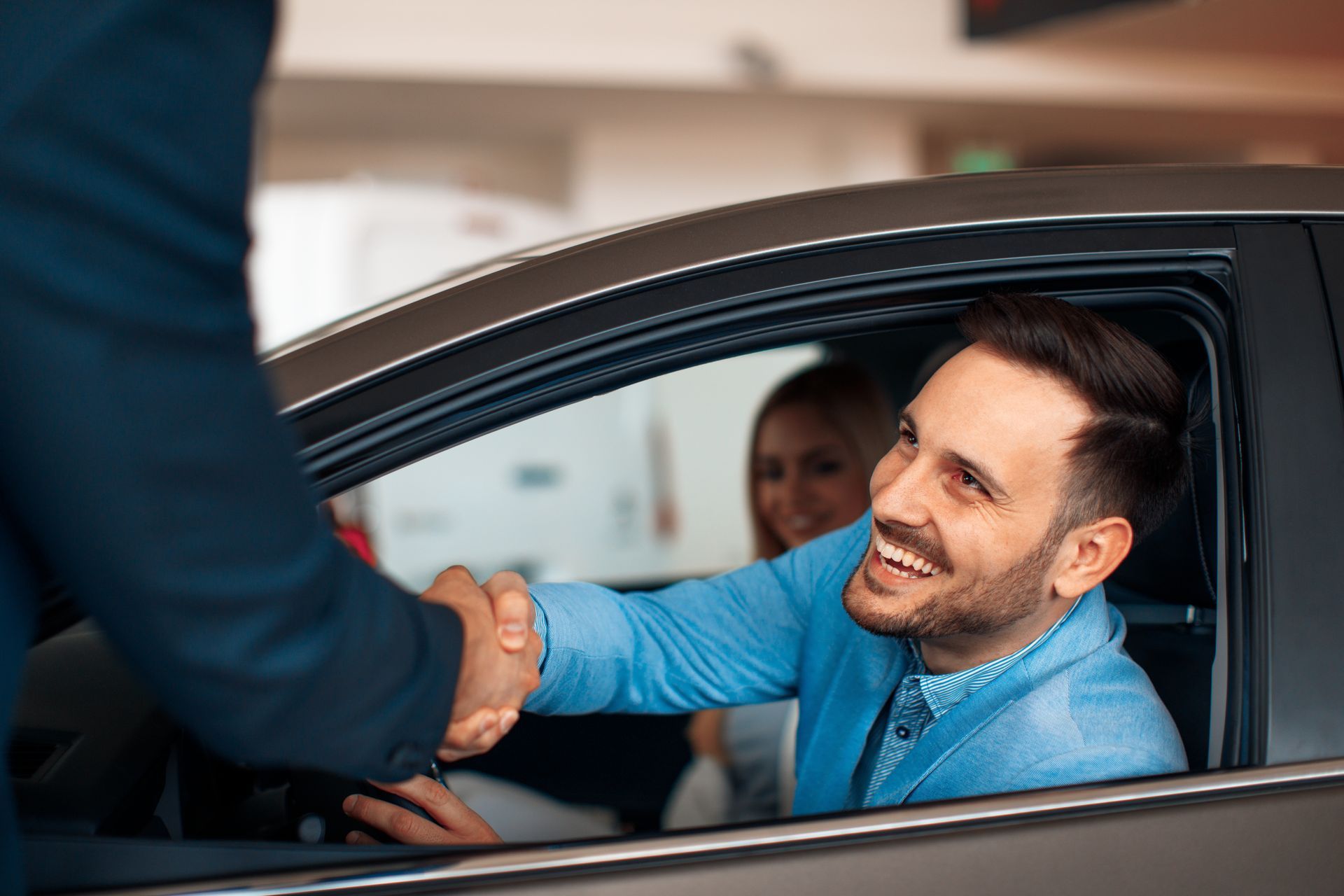 Man in new car shakes hands with a salesperson, woman in the backseat smiles.