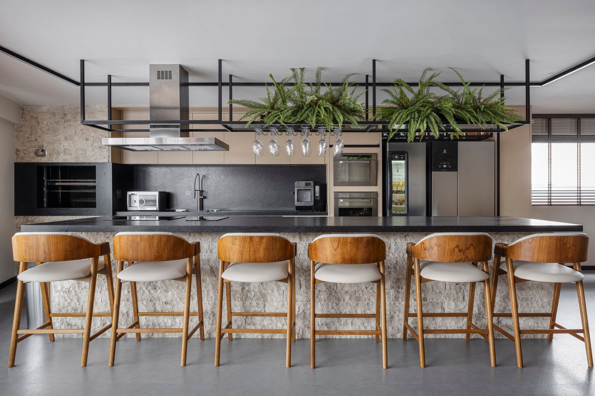 Cozinha com balcão tipo ilha, bancos de madeira, bancada de granito, eletrodomésticos de aço inoxidável e plantas suspensas.