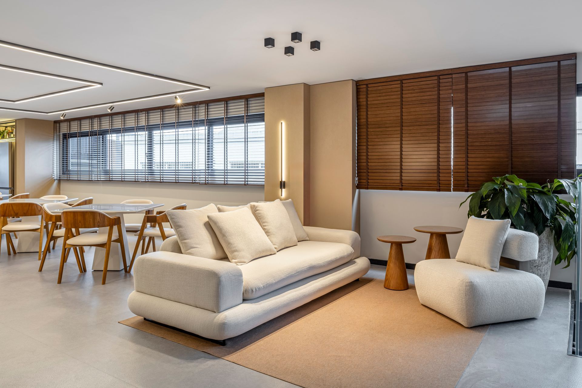 Sala de estar com sofá bege, poltrona, mesas laterais de madeira e área de jantar com mesa e cadeiras.