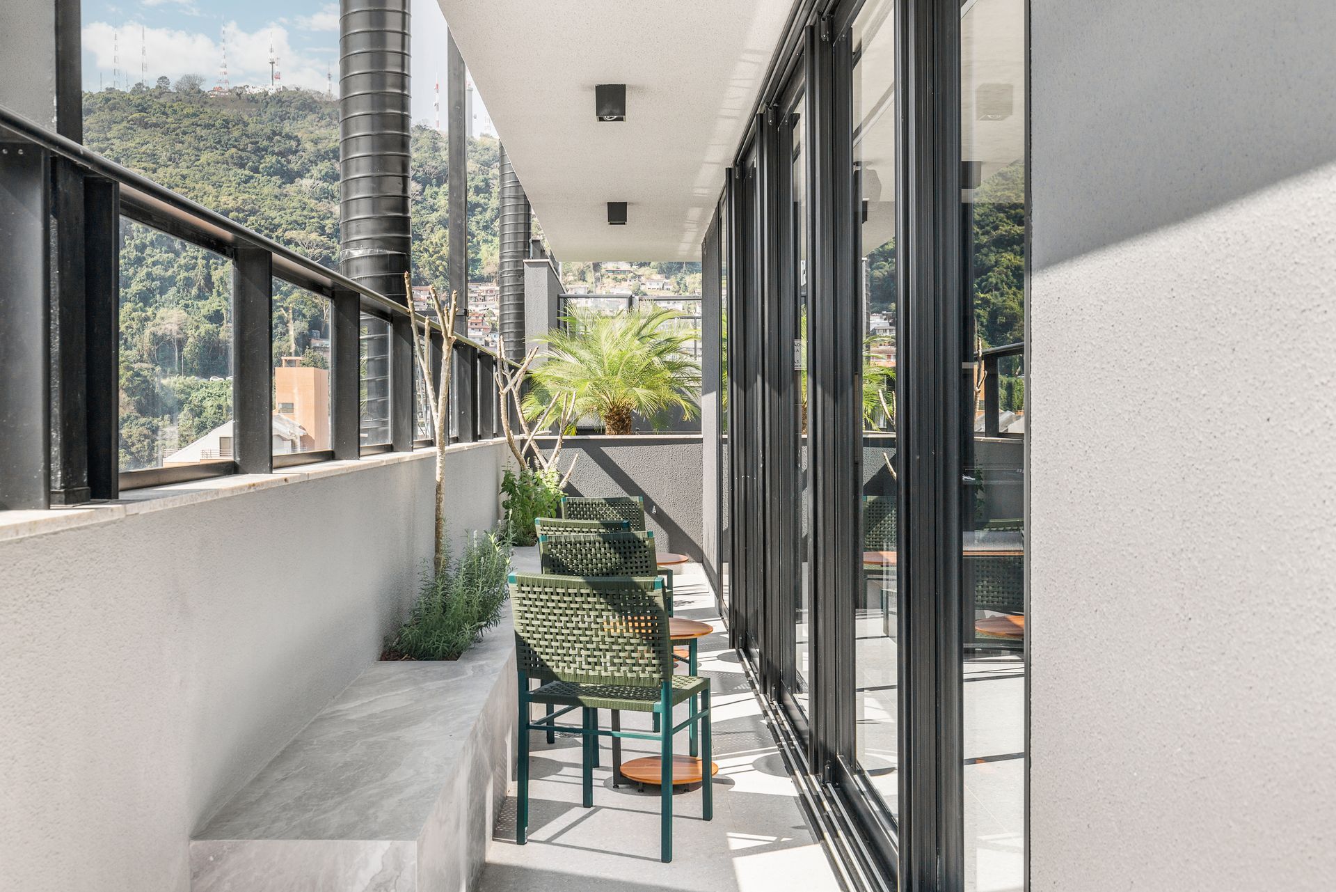 Varanda com portas de vidro, uma cadeira verde e um banco embutido, com vista para uma encosta verdejante.