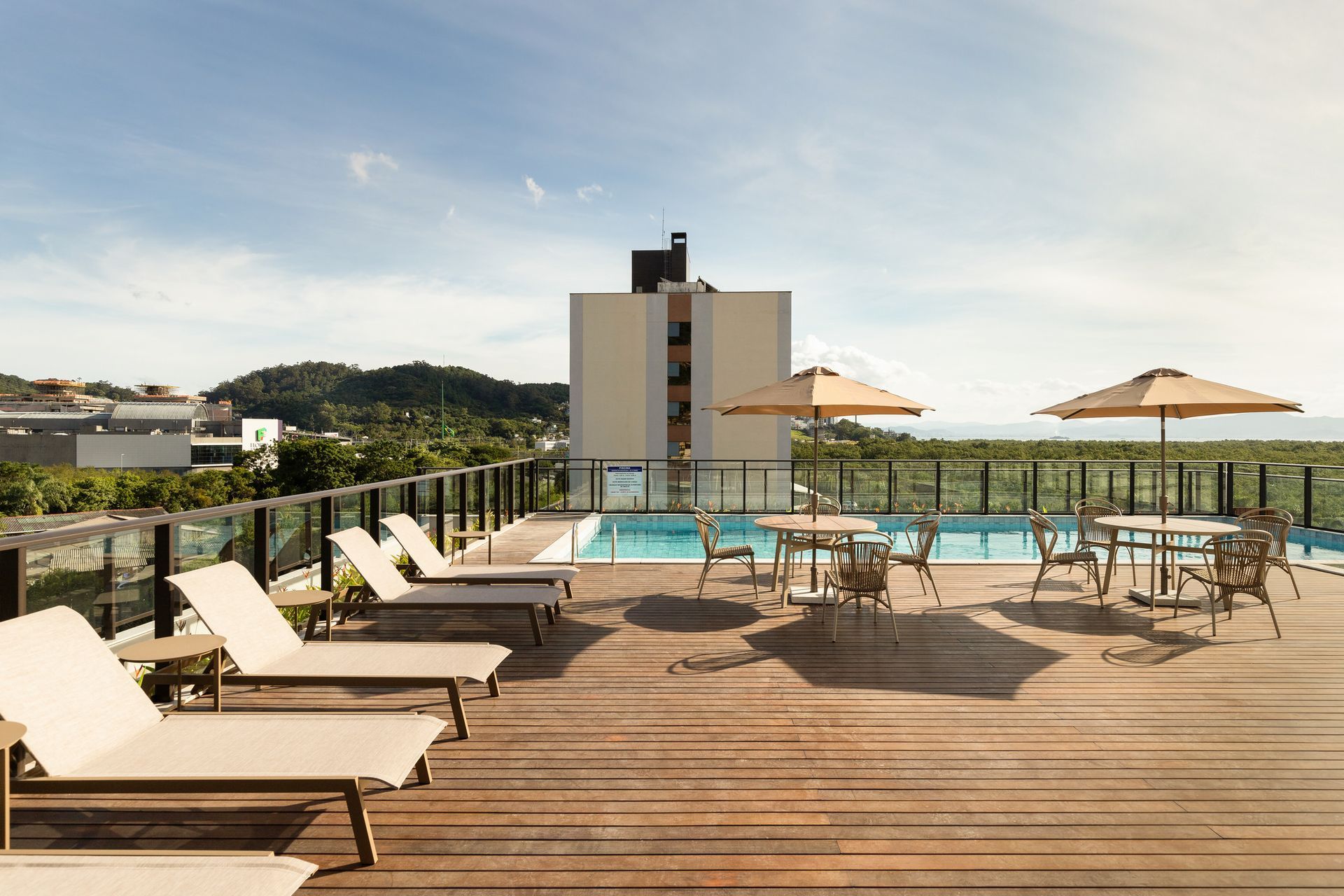 Piscina na cobertura com espreguiçadeiras, mesas, guarda-sóis e um prédio. Céu azul e vegetação ao fundo.