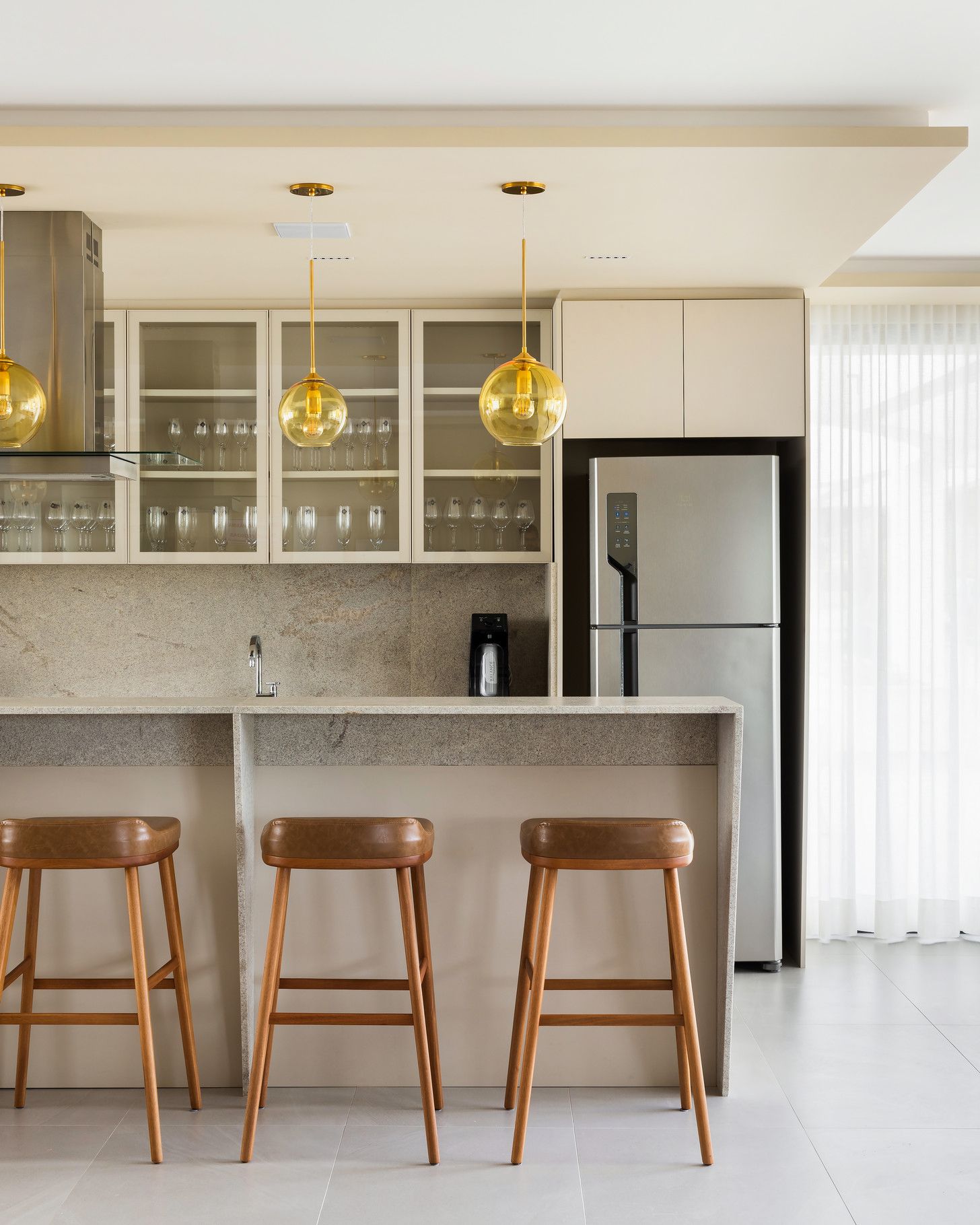 Cozinha moderna com bancos de bar, armários, geladeira e luminárias pendentes.