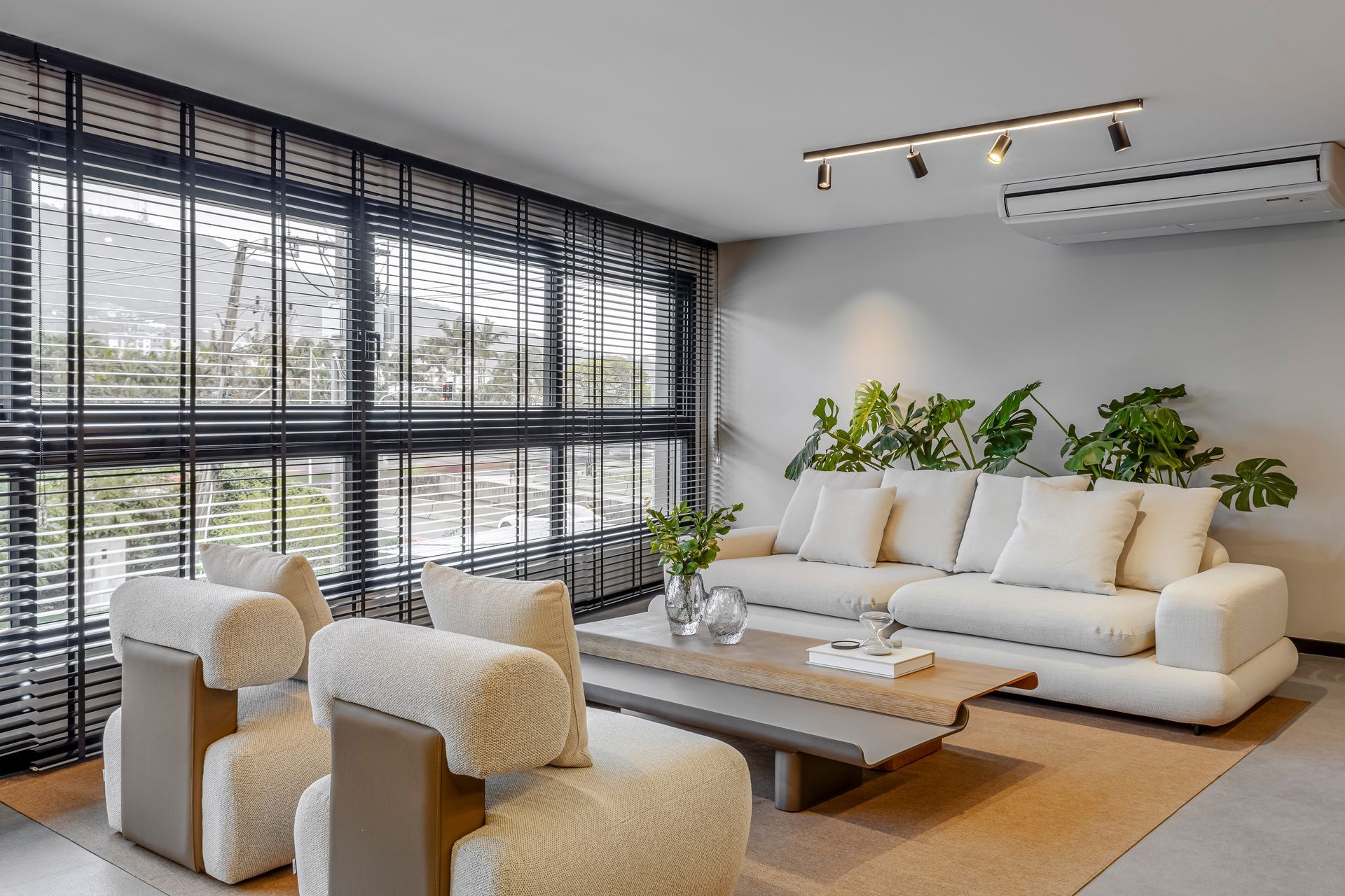 Sala de estar com sofá cor creme, poltronas, janela grande e plantas de interior.