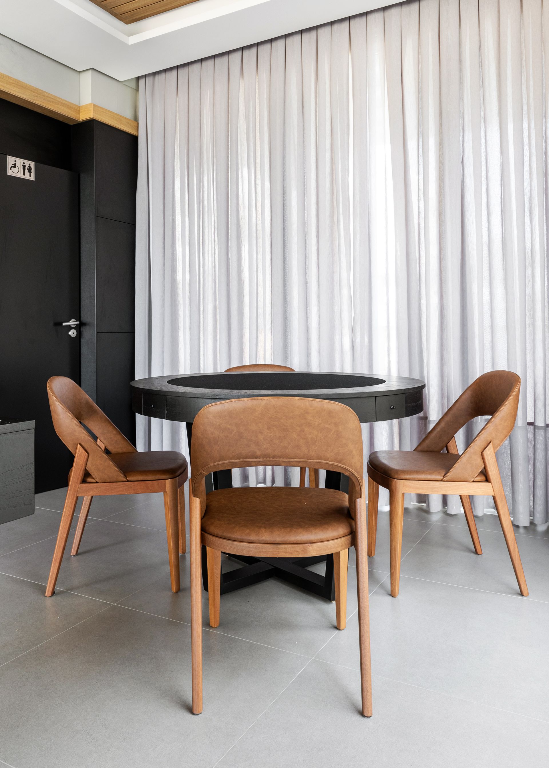 Quatro cadeiras marrons ao redor de uma mesa redonda escura, em frente a cortinas brancas, ao lado de uma porta preta.