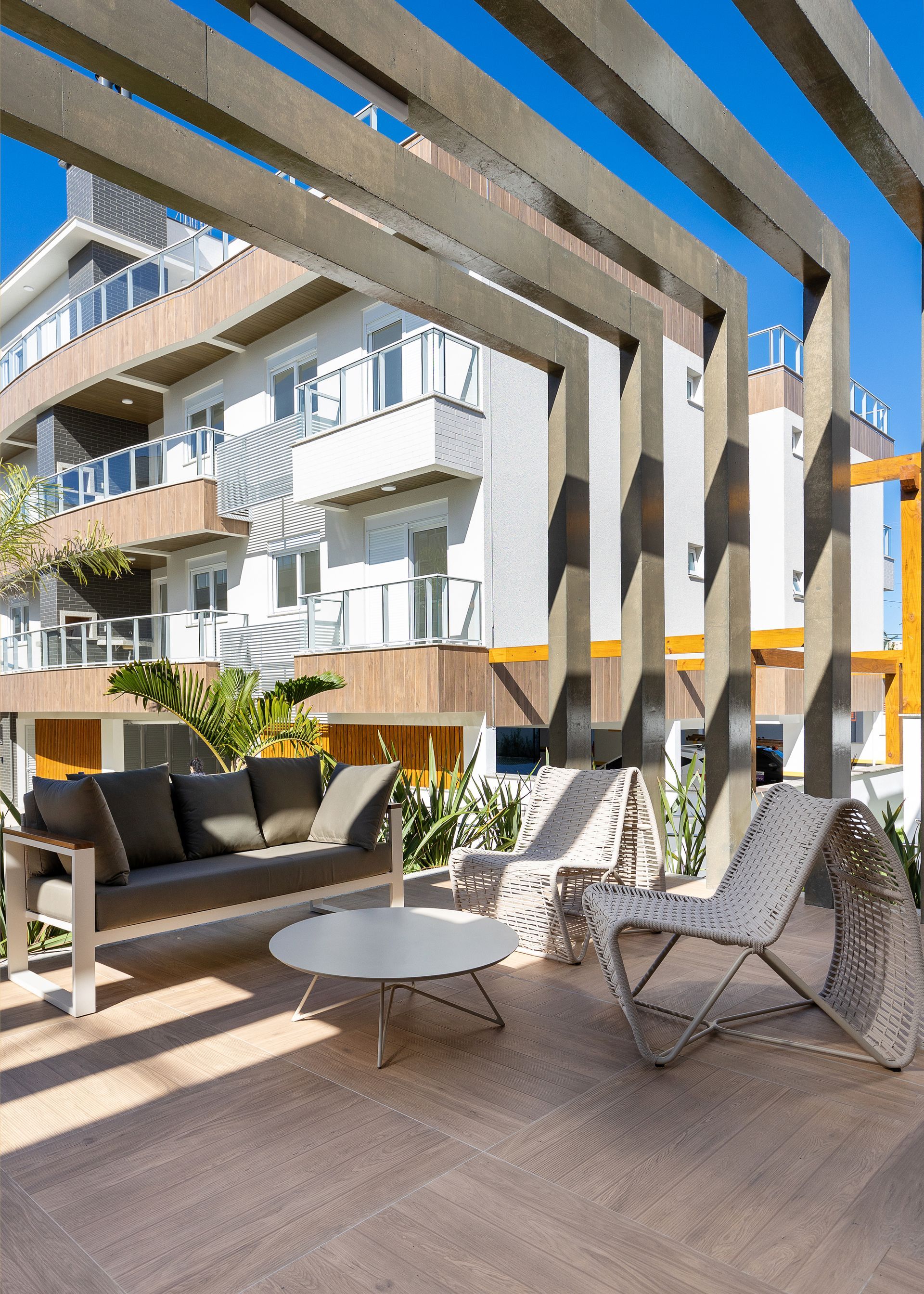Pátio externo com móveis modernos sob uma pérgola de madeira, ao lado de um prédio de vários andares.