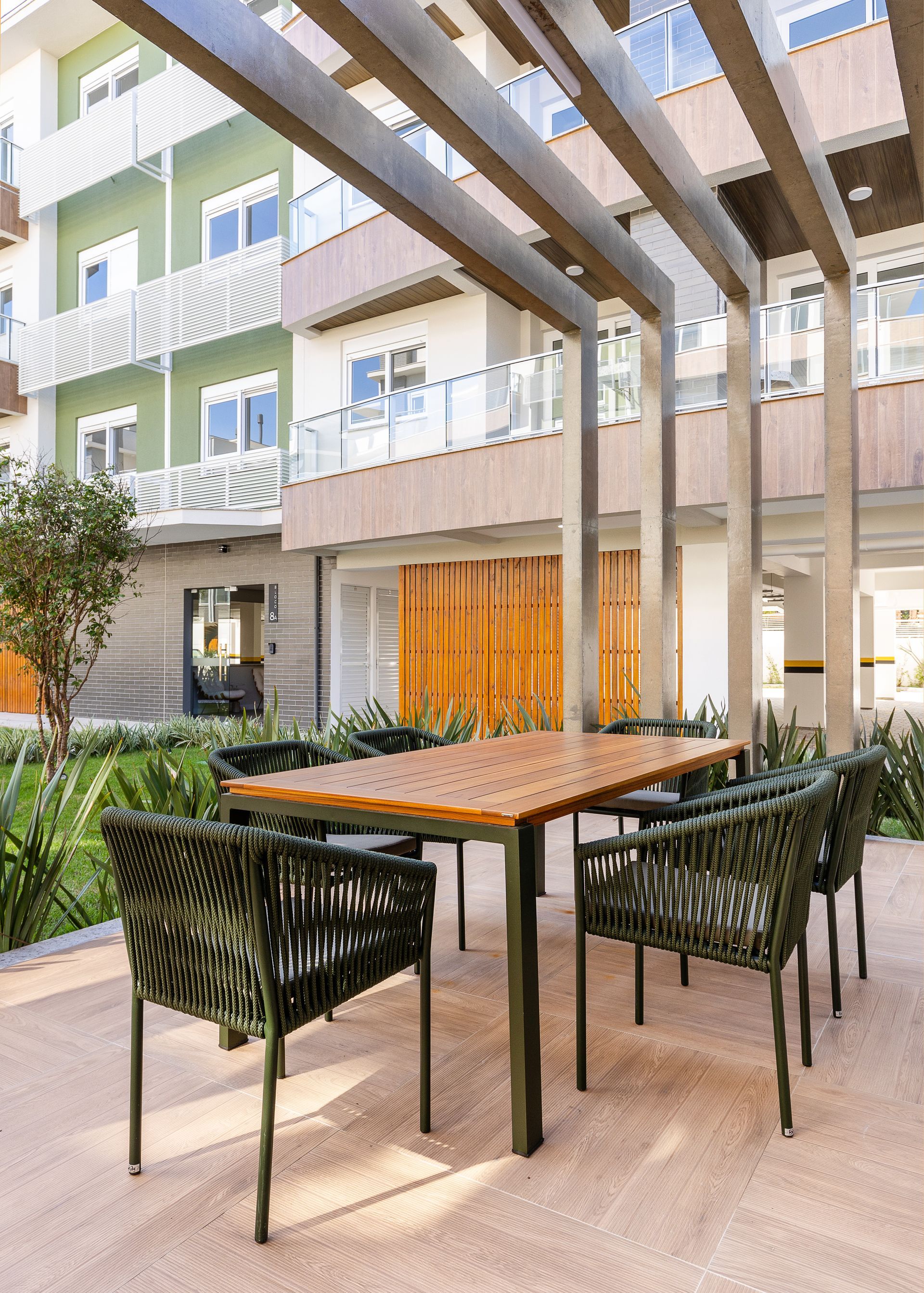 Conjunto de jantar ao ar livre sob uma pérgola, com cadeiras, mesa e um prédio ao fundo.