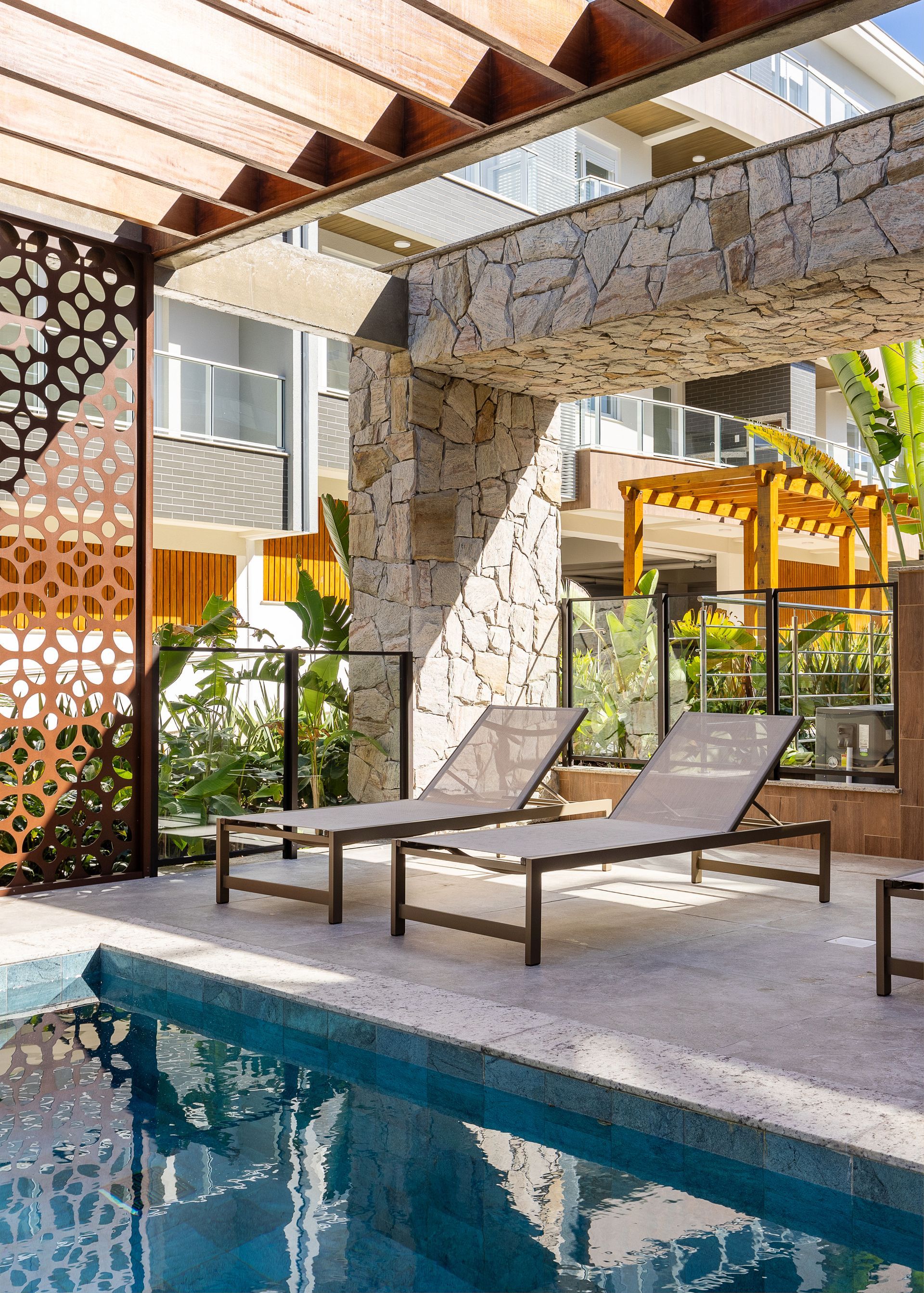 À beira da piscina com duas espreguiçadeiras, sob uma pérgola de pedra e madeira.