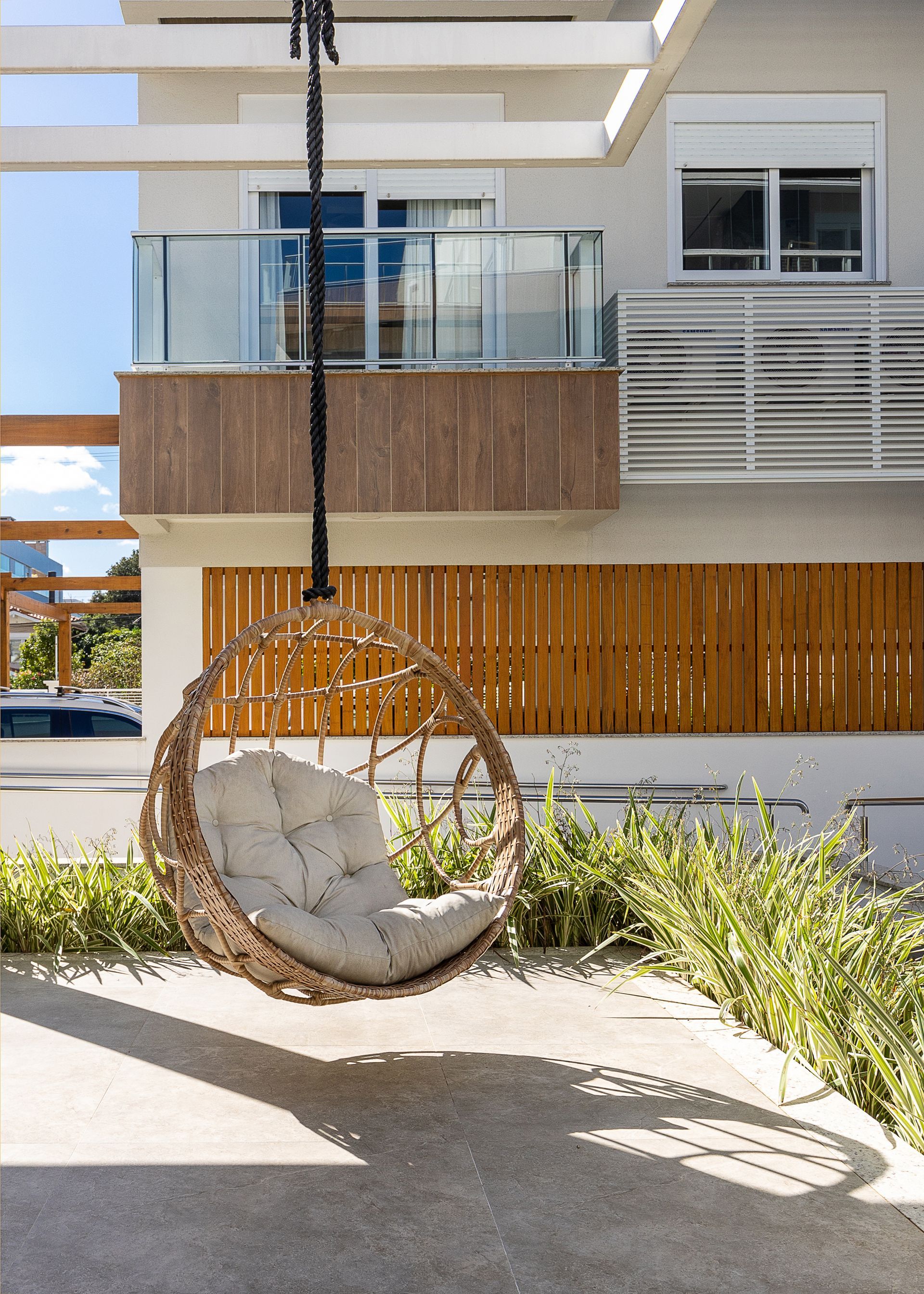 Cadeira suspensa de vime com almofada, em frente a um edifício moderno com detalhes em madeira.