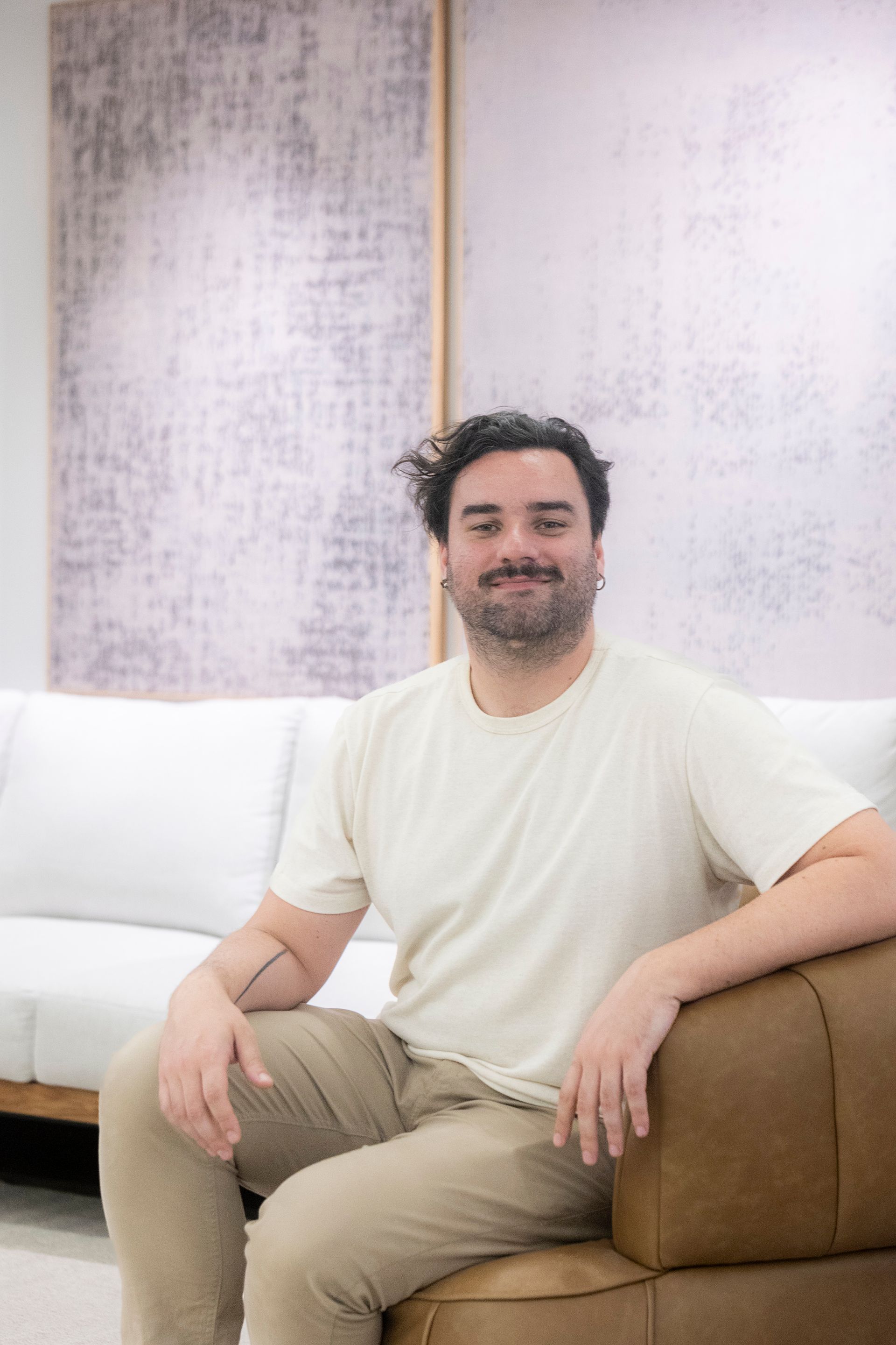 Homem de cabelos escuros, sentado em um sofá marrom em uma sala de cor clara, sorrindo.