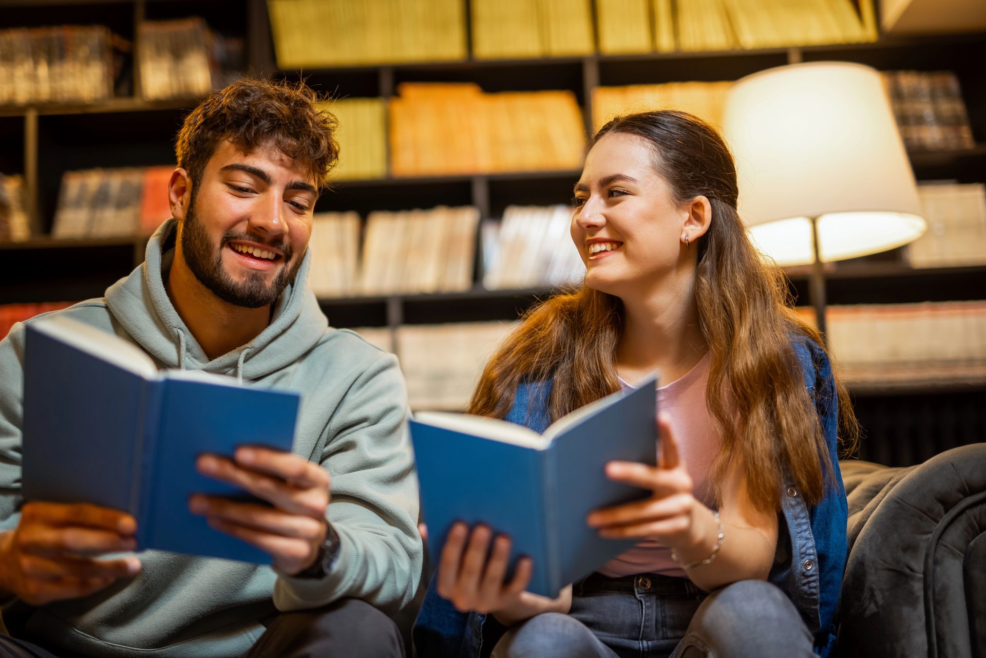 Un uomo e una donna sorridono mentre leggono un libro in una biblioteca, sullo sfondo ci sono degli scaffali.