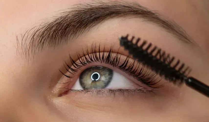 Primo piano di un occhio, mentre si applica il mascara con un pennello. L'occhio è verde con lunghe ciglia.