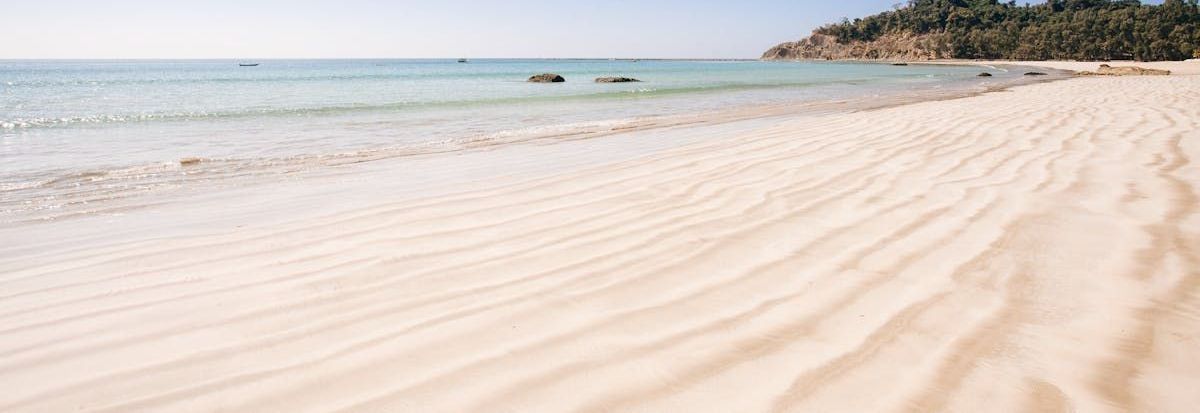 Sandy beach with gentle waves, clear blue water, and a rocky shoreline under a bright sky