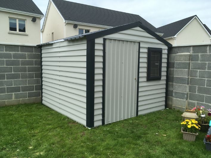 A white shed with a black trim is in a backyard next to a brick wall.