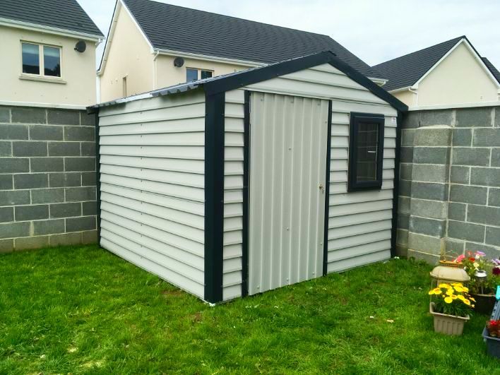 A white shed is sitting in the grass in front of a brick wall.
