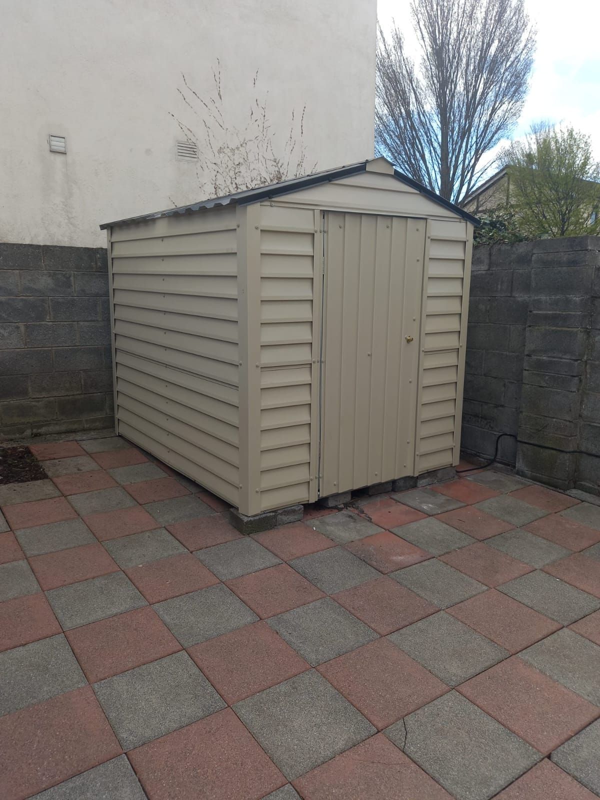 A small white shed is sitting on a tiled patio in front of a building.