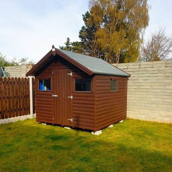 A wooden shed with a green roof sits in a lush green yard