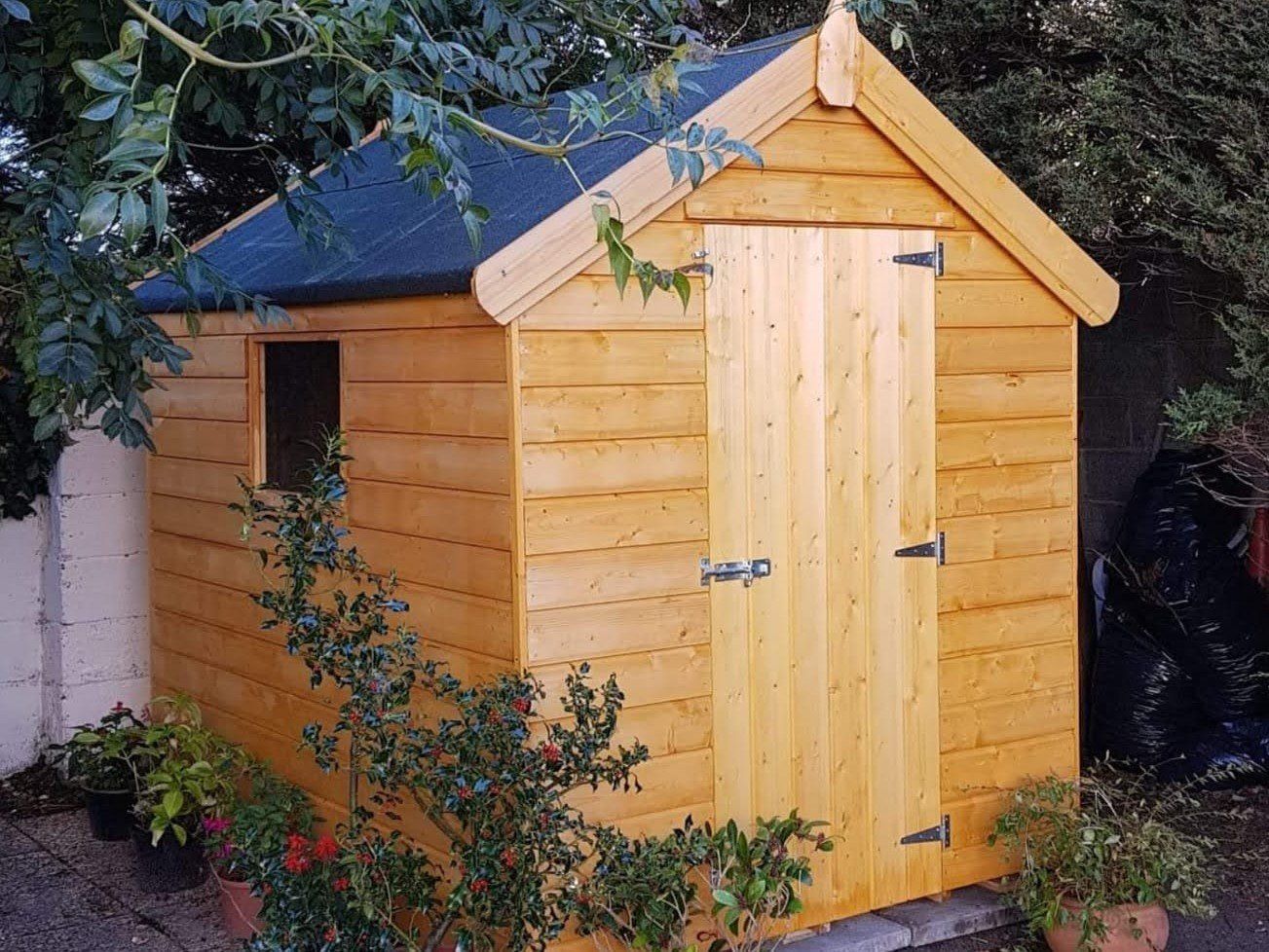 A wooden shed with a black roof is surrounded by trees and potted plants.