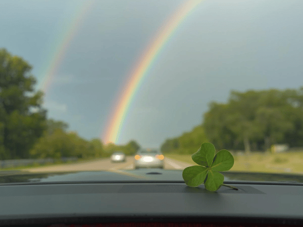 View from a car dashboard with a four-leaf clover and a double rainbow over the road ahead.