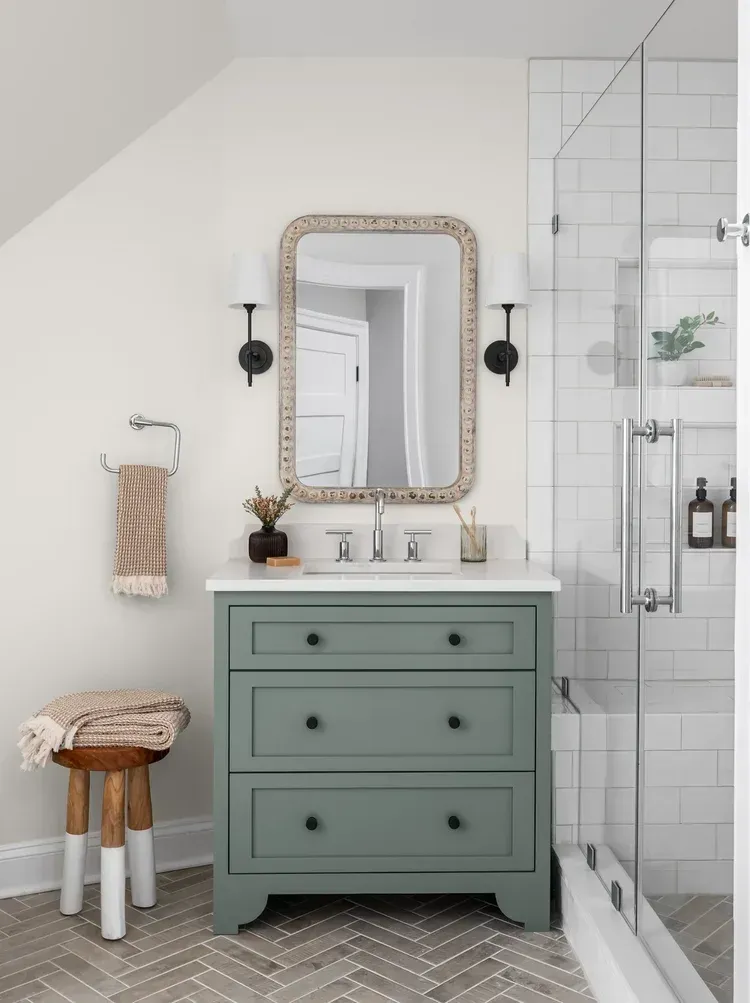 Bathroom with green vanity, white countertop, ornate mirror, glass shower, and herringbone floor.