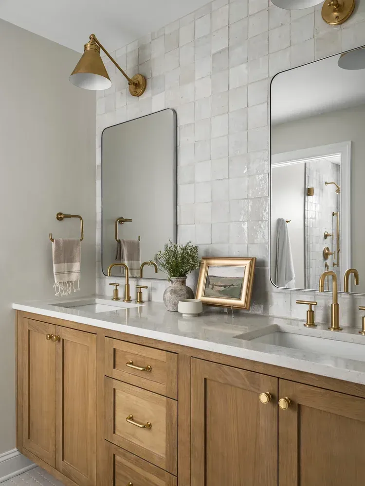 Bathroom with light wood vanity, marble countertop, gold fixtures, square tile wall, and two mirrors.