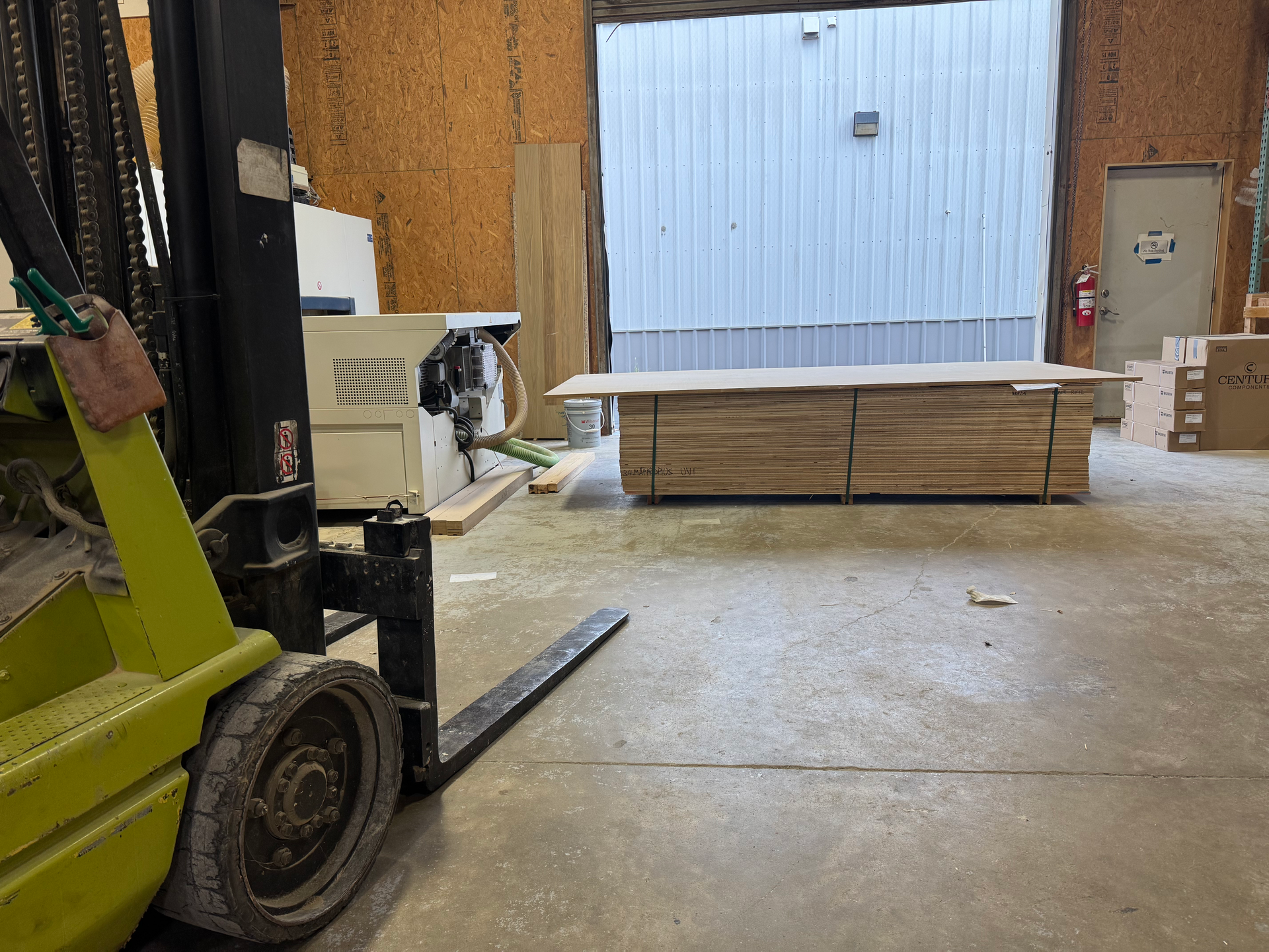 A green forklift with black forks inside a warehouse. Wood planks are stacked in the background near the entrance.