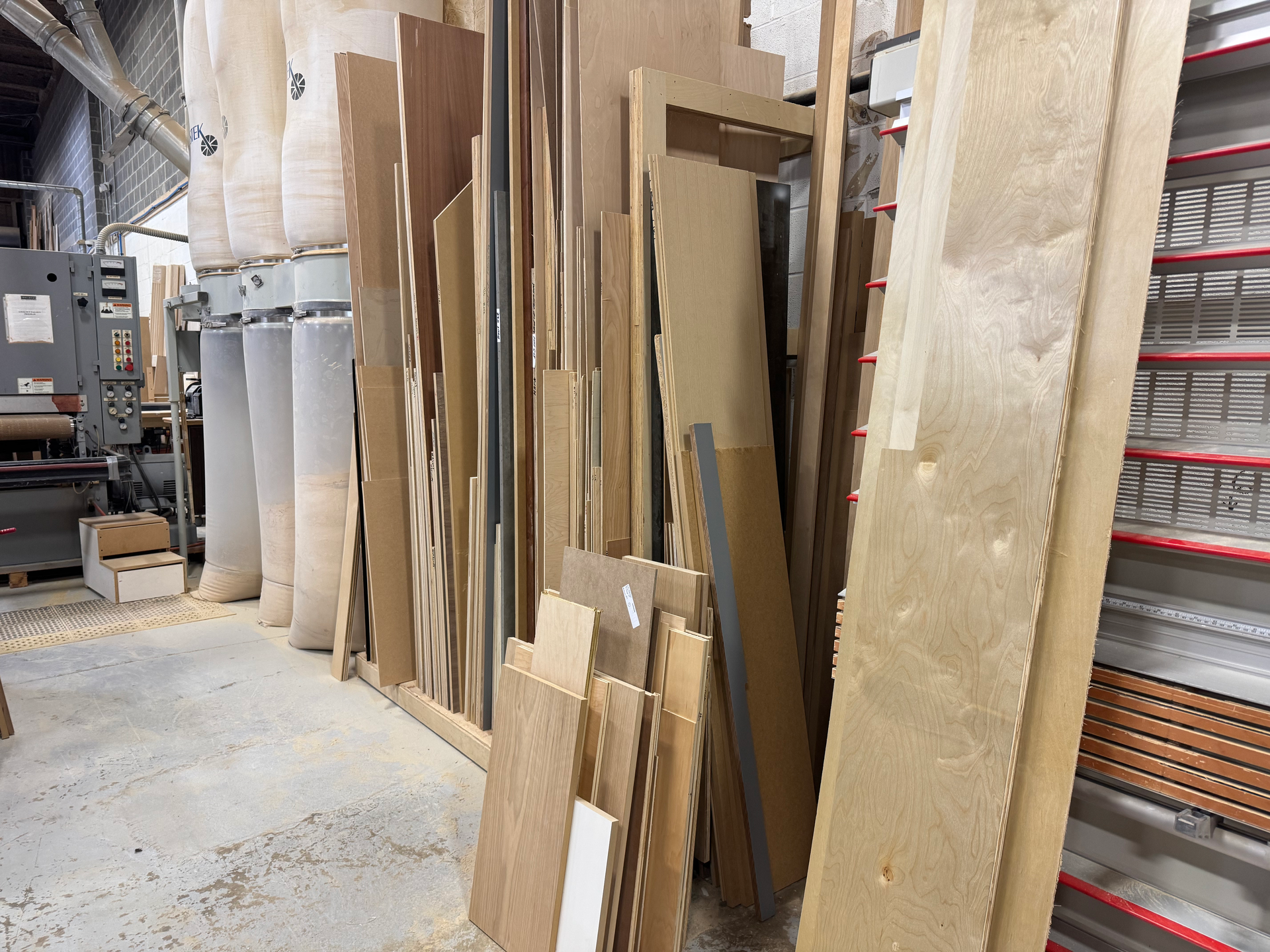 Wood planks and panels stored in a woodworking shop, varying in size and color, next to machinery.