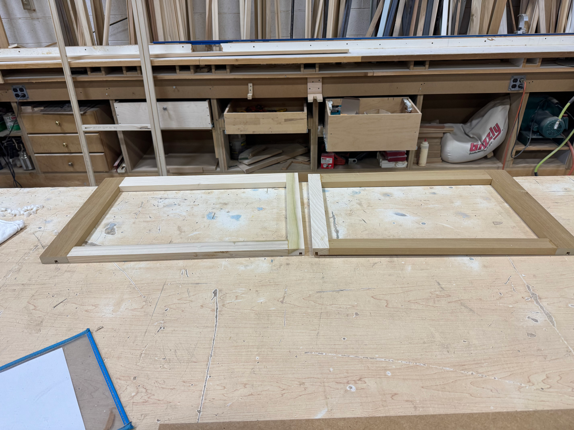Two wooden frames are on a workshop floor. Shelves with materials and tools are in the background.