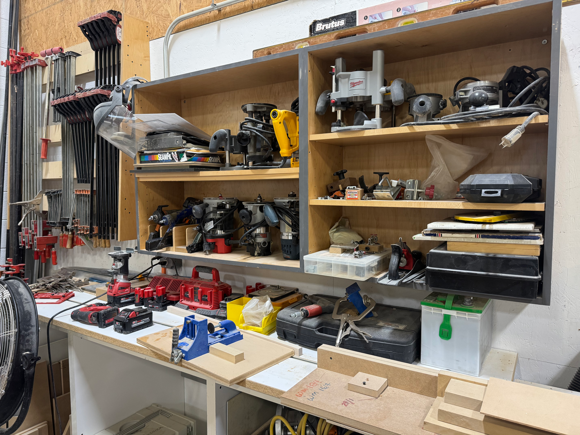 Workshop with tools on a workbench, shelves filled with machinery, and clamps hanging above.