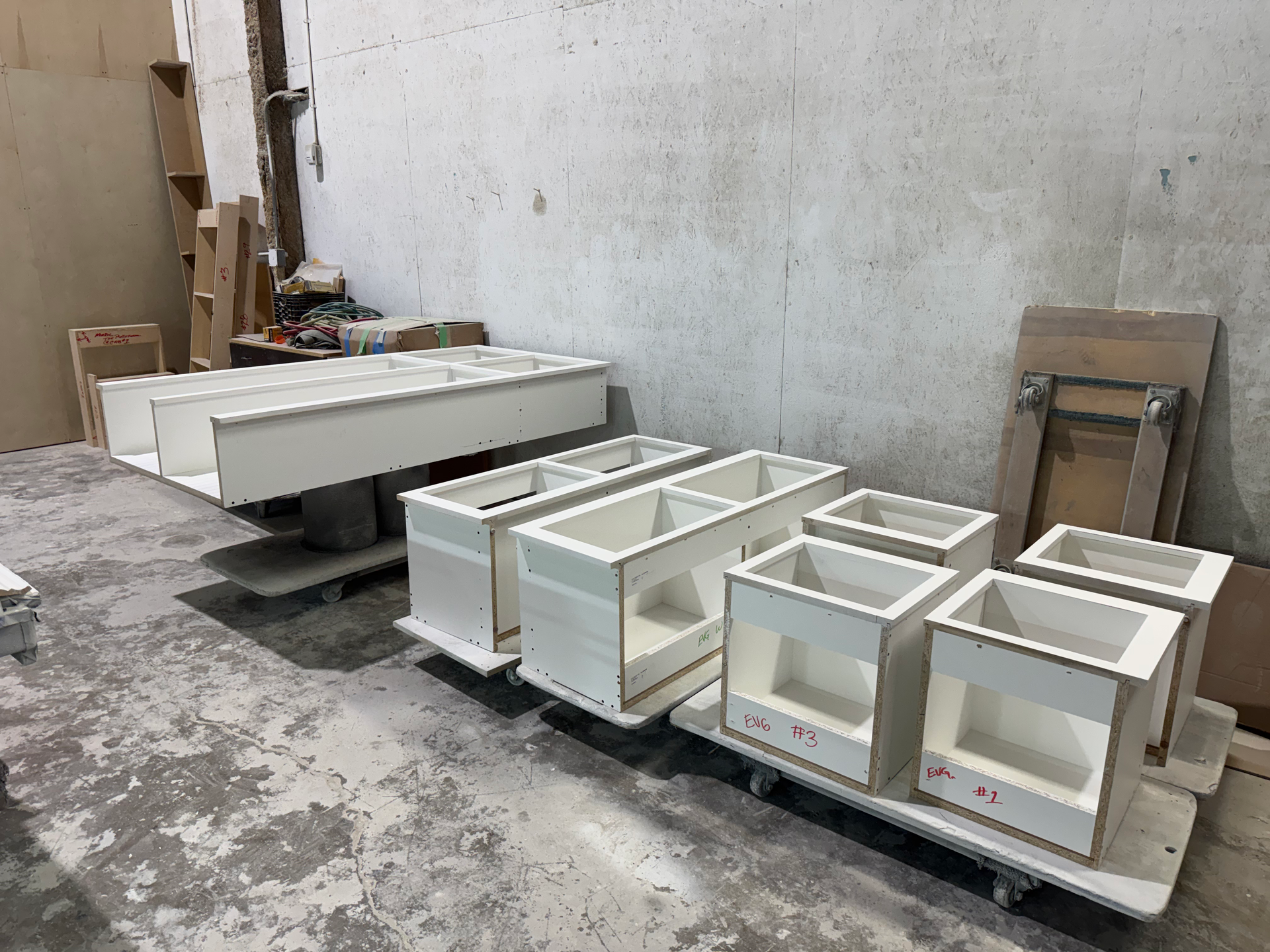 White cabinetry components in various stages of assembly, in a workshop with a concrete floor.
