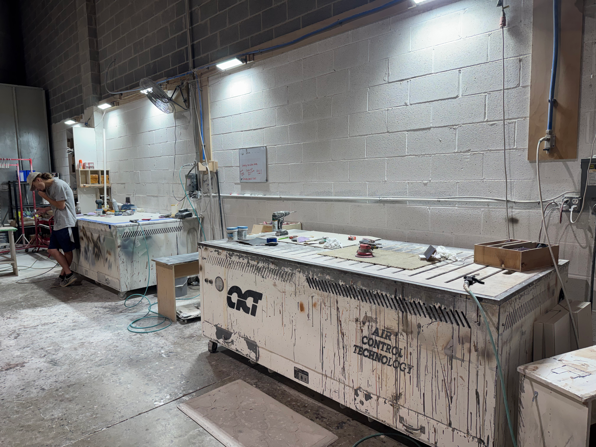 Workshop with two cluttered workbenches, a person working on one, and overhead lighting.