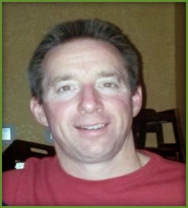 Man with short brown hair wearing a red shirt smiling. Background is out of focus.