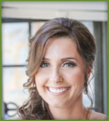 Woman with brown hair styled up, smiling broadly. Wearing makeup, against a blurred background.