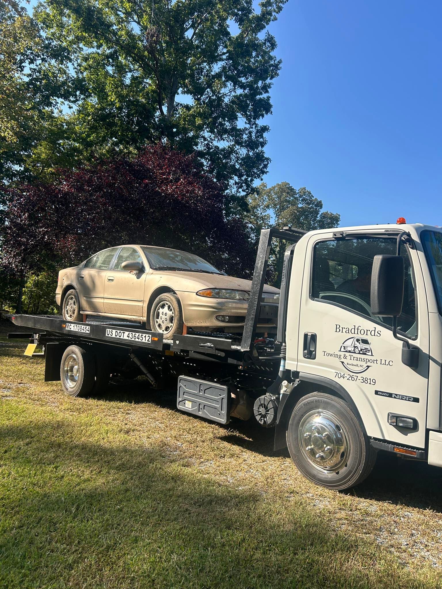 Tow truck carrying a gold sedan on a grassy roadside under a clear blue sky