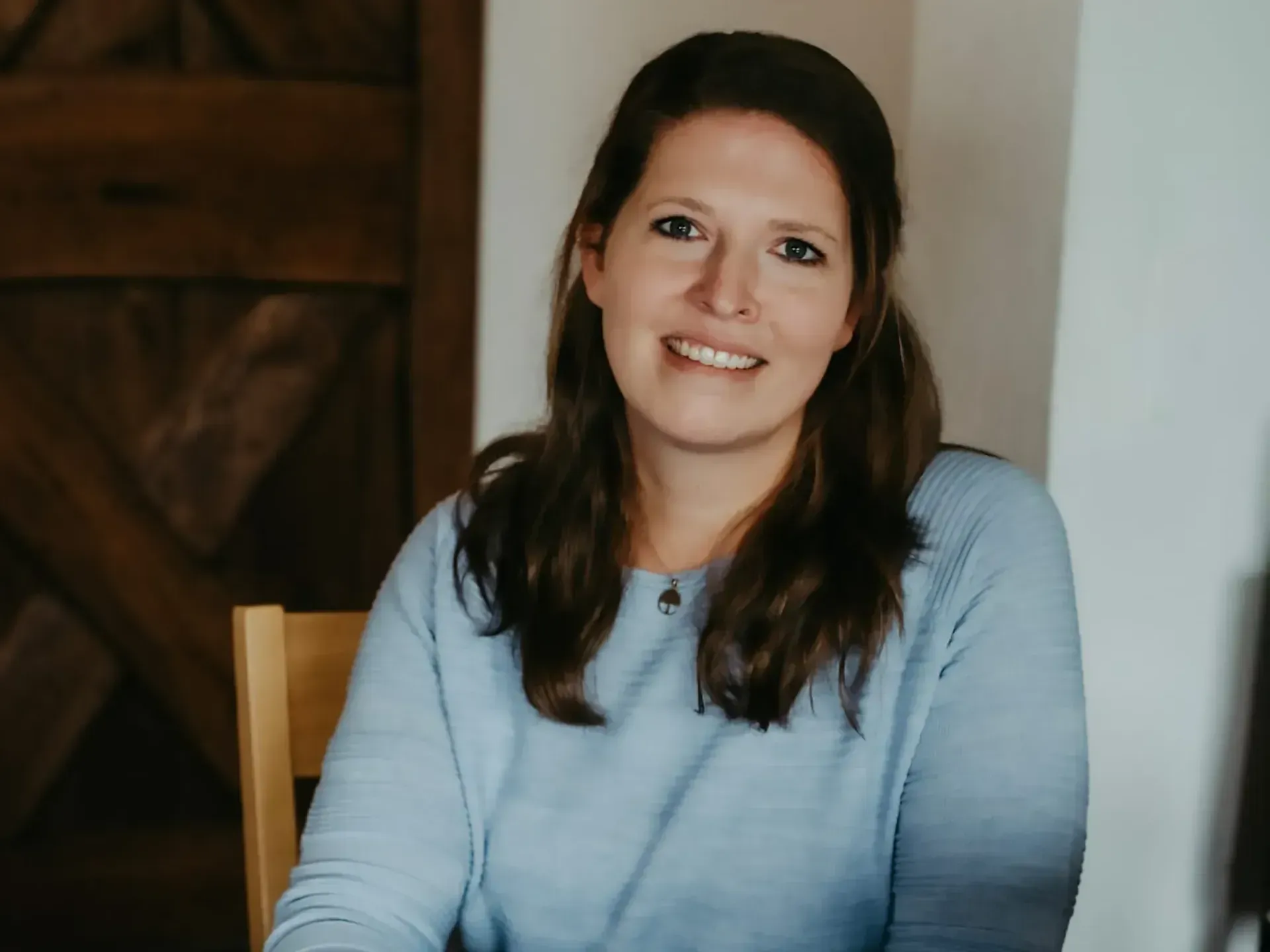 Julia in einem blauen Pullover sitzt auf einem Stuhl und lächelt in die Kamera.