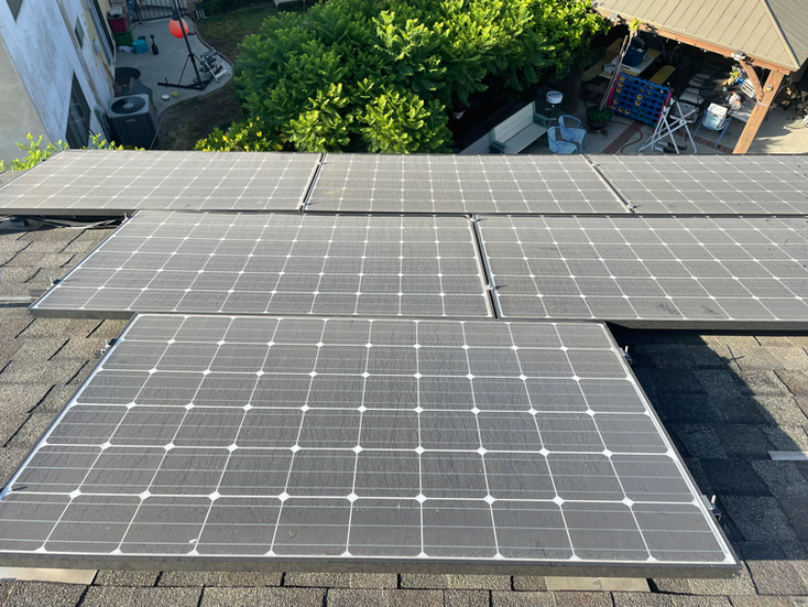 Solar panels installed on a dark gray shingled roof against a background of green trees and a building.