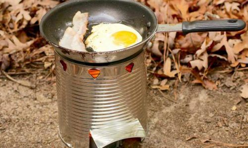 A frying pan filled with eggs and bacon is cooking on a tin can stove.