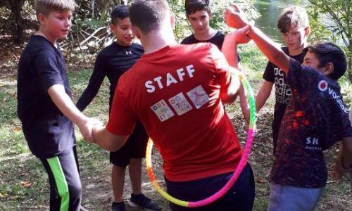 A man in a red staff shirt is holding a hula hoop in a circle of children.