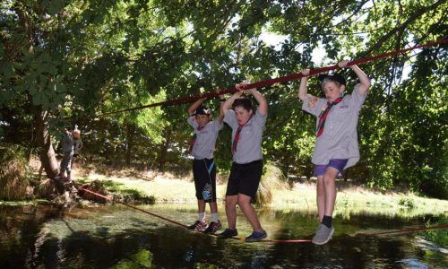 Three boy scouts are crossing a river on a rope bridge.