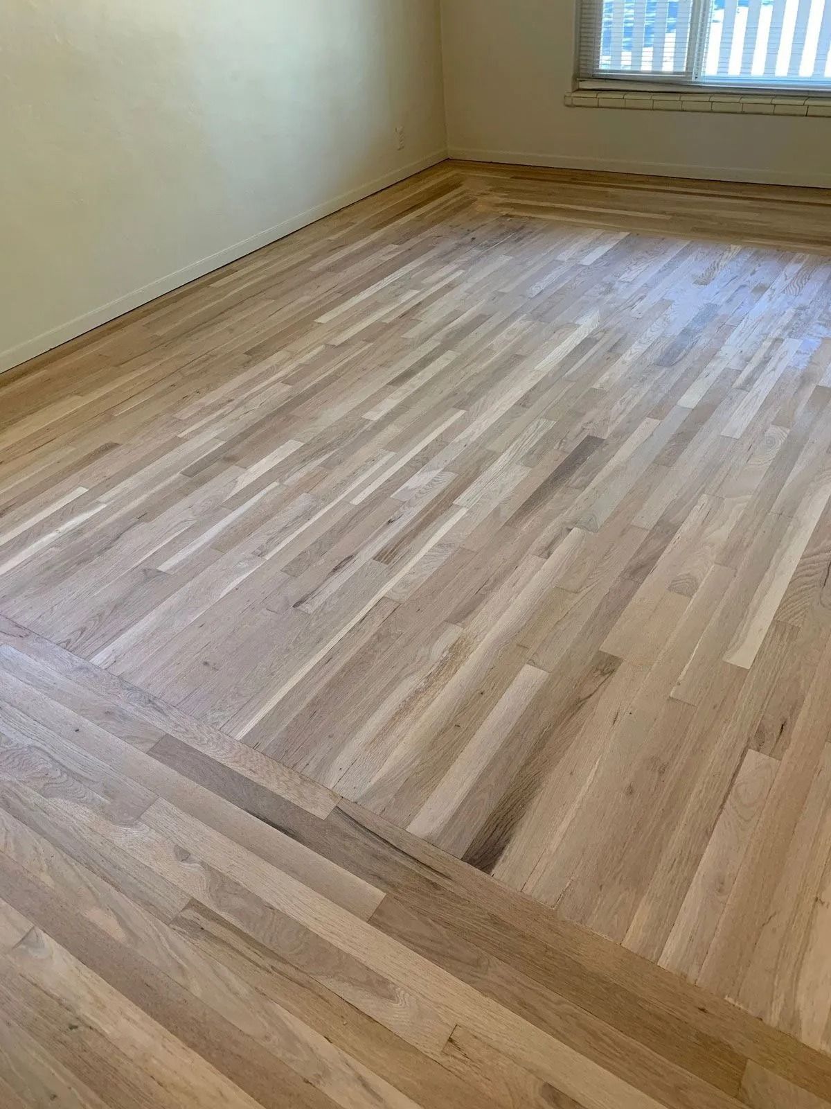 Light-colored wooden floor in herringbone pattern, with a white wall and window in the background.