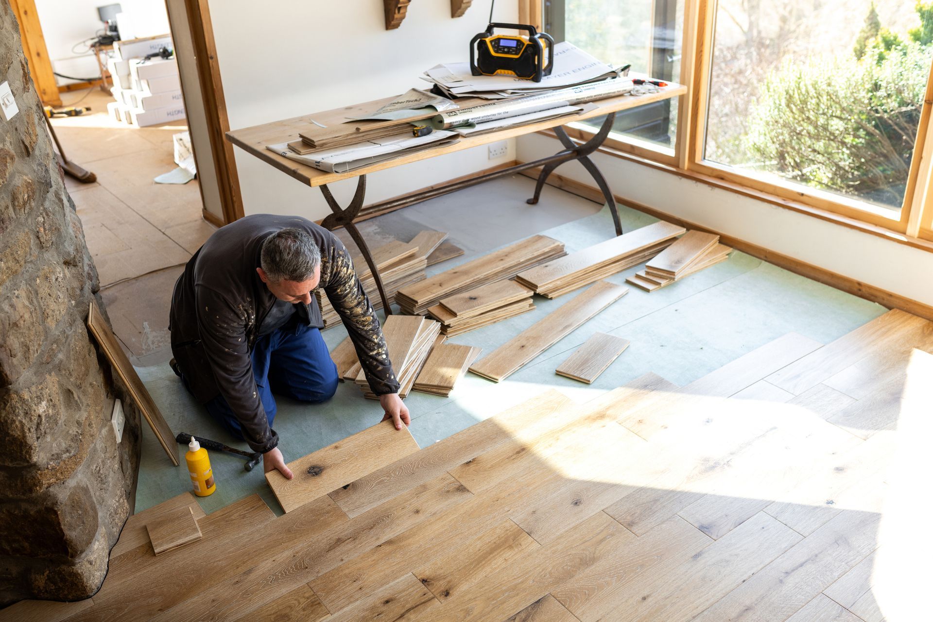 Man installing hardwood floor planks, kneeling on the floor near window. Beige planks, blue underlayment, and natural light.