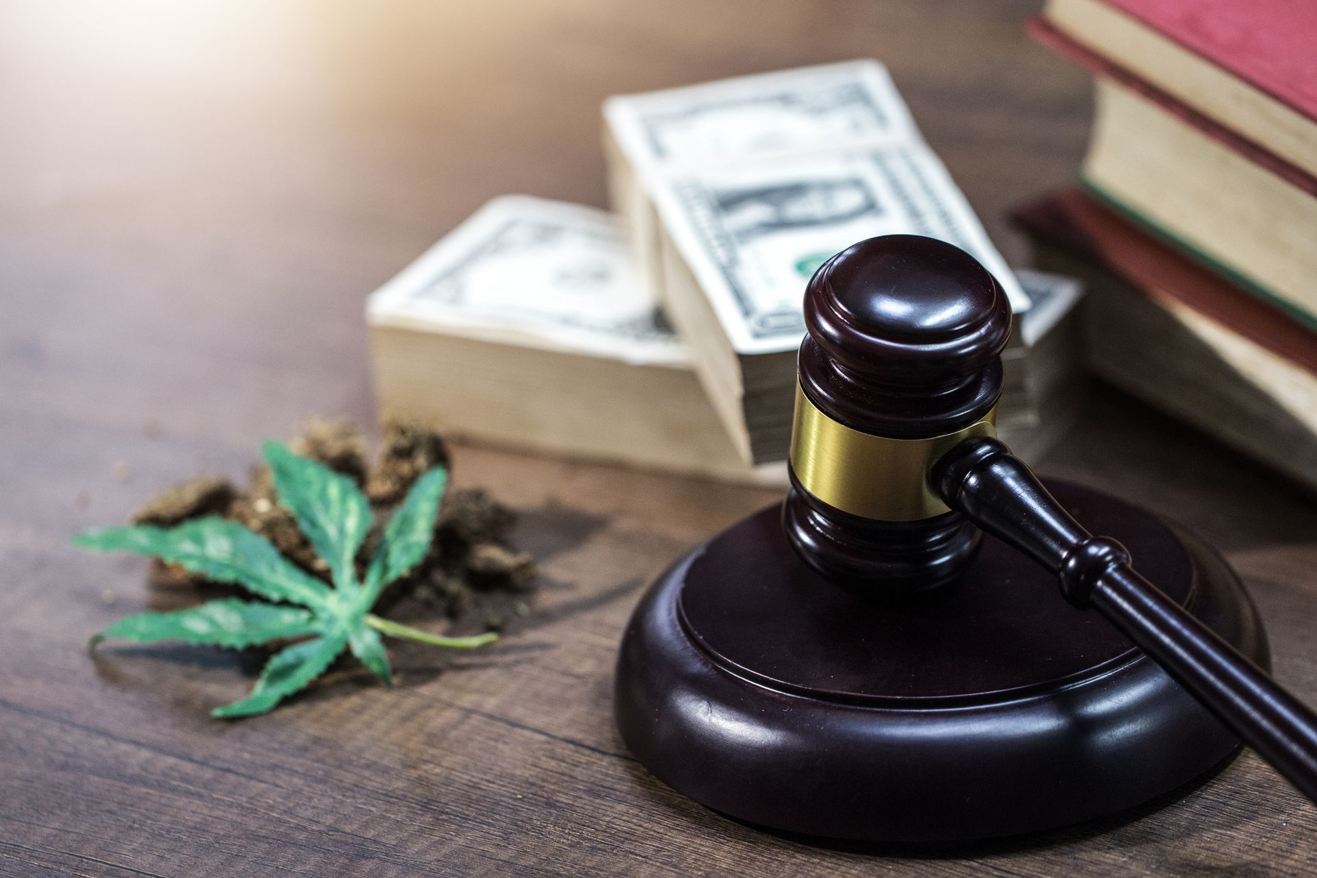 Judge's gavel, cannabis leaf, dried buds, money stacks, and red books on a table.