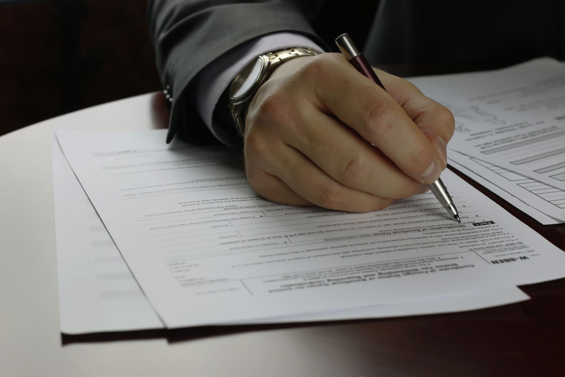 A person's hand in a business suit writing on a stack of papers, likely at a desk.
