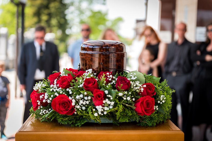 A funeral urn sits atop a wooden surface surrounded by a decorative wreath of red roses and white baby's breath.