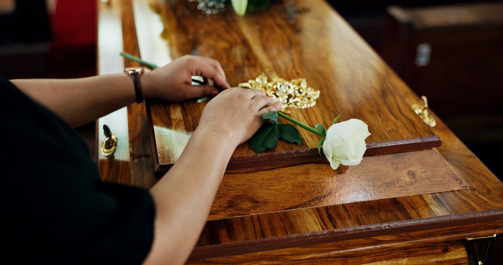 A person’s hands rest gently on a wooden casket, holding a single white rose near a small gold floral arrangement.